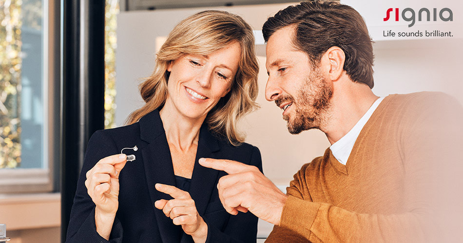blonde_haired_woman_wearing_black_blazer_smiles_and_holds_signia_hearing_aid_from_pure_sound_lititz_location_while_brown_haired_bearded_man_with_orange_brown_sweater_points_and_looks_at_hearing_aid