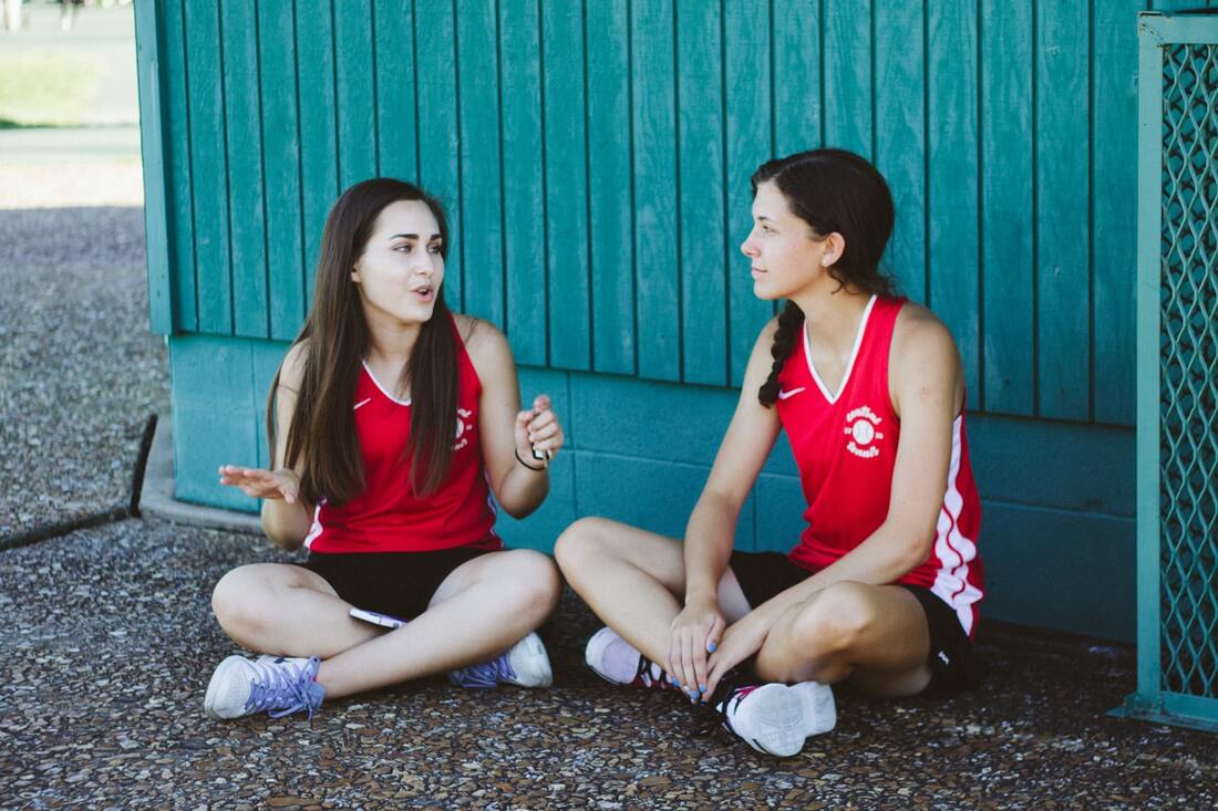 Two girls in red and white tank tops, black shorts, and white sneakers sit on the ground and talk by a turquoise building.