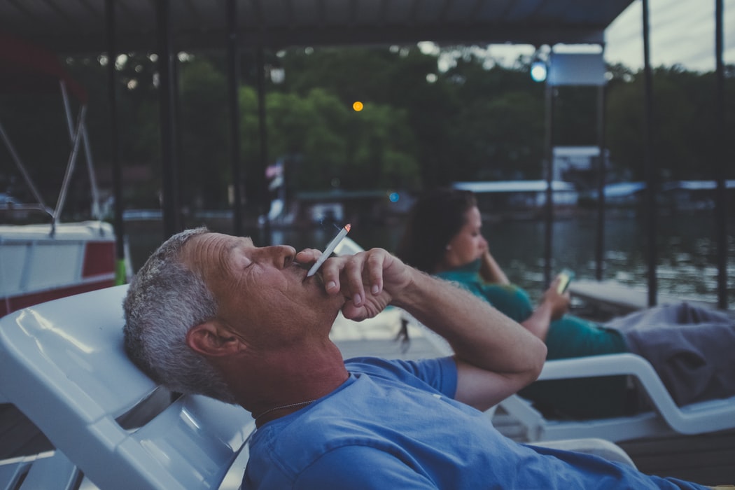 A man lays on a white poolside lounge chair and smokes while leaning his head back.