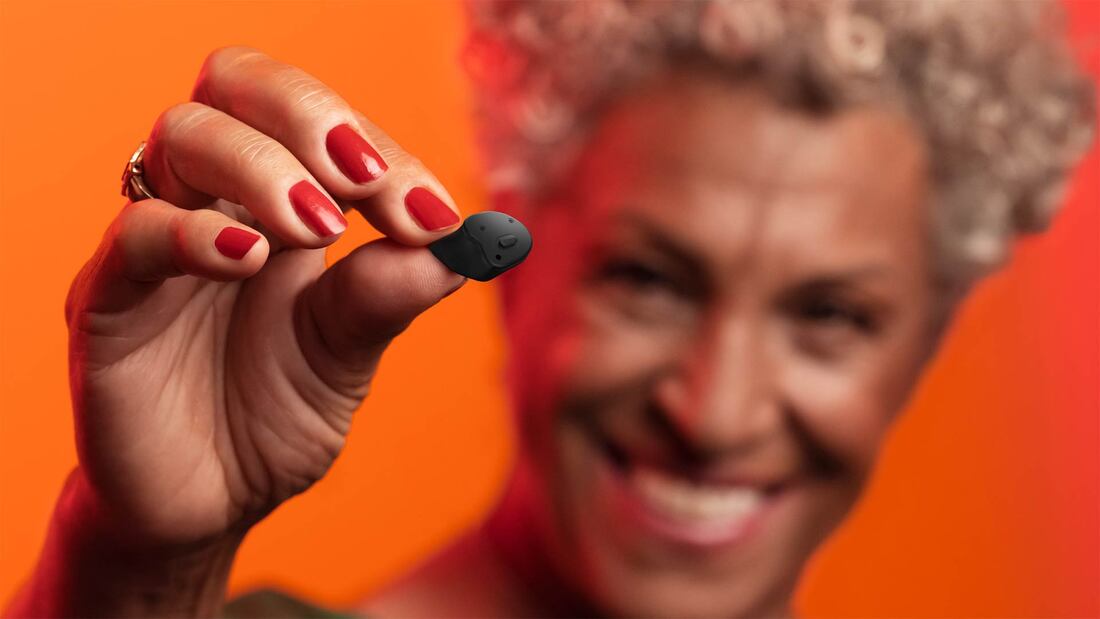 A woman with painted red nails and gray hair holds a small in-the-ear hearing aid.