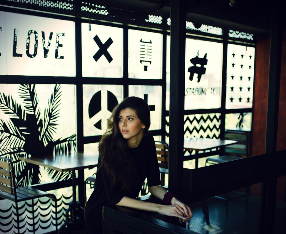 A woman with brown hair wears dark clothes and stands inside a cafe with windows decorated with images and words in black.