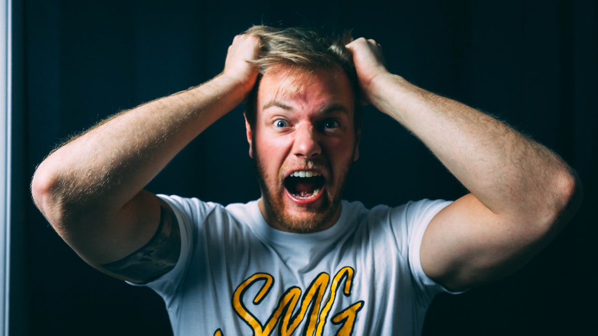 A man in a white t-shirt with yellow text places his hands on his head and screams in frustration at annoying noises.