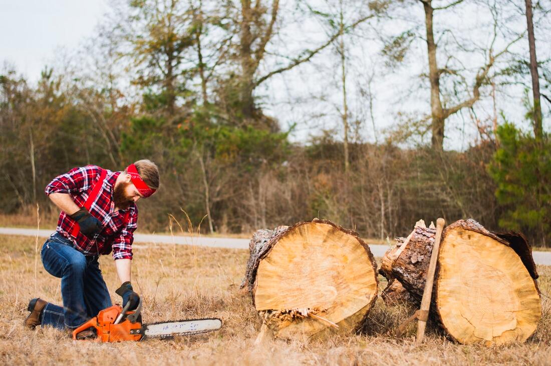 bearded_pennsylvania_man_wearing_red_flannel_shirt_blue_jeans_with_red_suspenders_red_bandana_blue_and_black_gloves_brown_boots_holds_orange_chainsaw_while_kneeling_by_cut_tree_trunk_has_hearing_loss