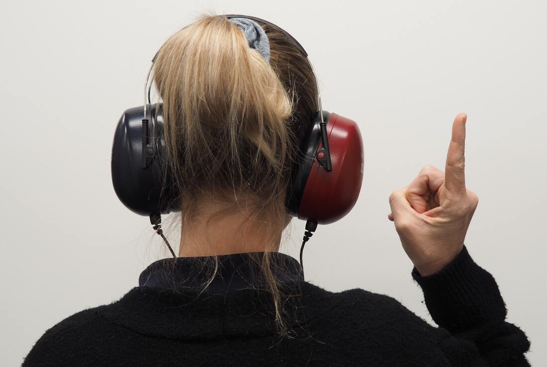 blonde_haired_woman_with_black_sweater_gets_hearing_test_wears_black_and_red_headphones_while_pointing_finger_up_at_pure_sound_hearing_strasburg_office