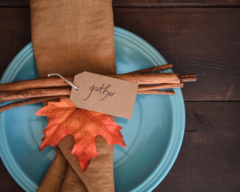 blue_plate_brown_cloth_napkin_cinnamon_sticks_red_orange_leaf_tag_with_gather_written_on_it_for_Thanksgiving