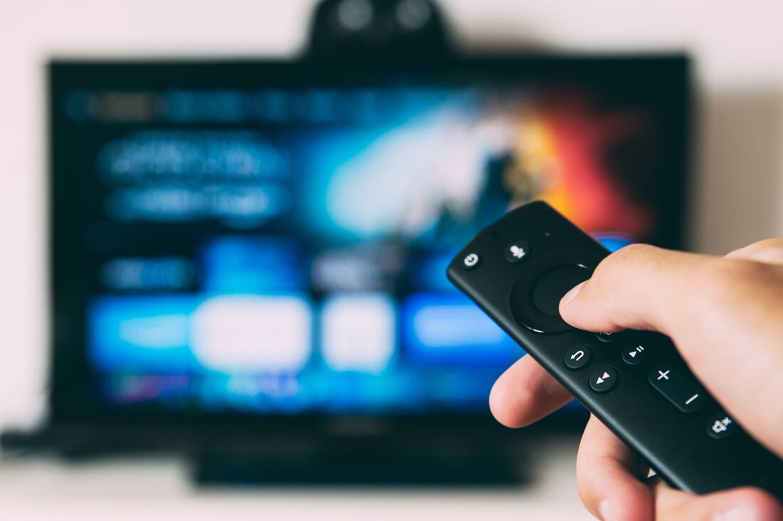 A T.V. in the background is out of focus, while a hand holds a black TV remote control with Bluetooth features at the forefront.