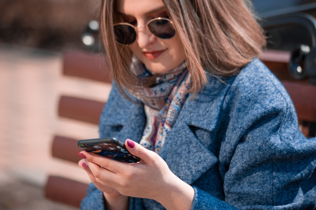 brown_haired_woman_with_copper-rimmed_sunglasses_black_tinted_lenses_wearing_blue_coat_pink_and_blue_scarf_holds_smartphone_with_hot_pink_painted_nails_while_sitting_on_bench_uses_hearing_aid_app