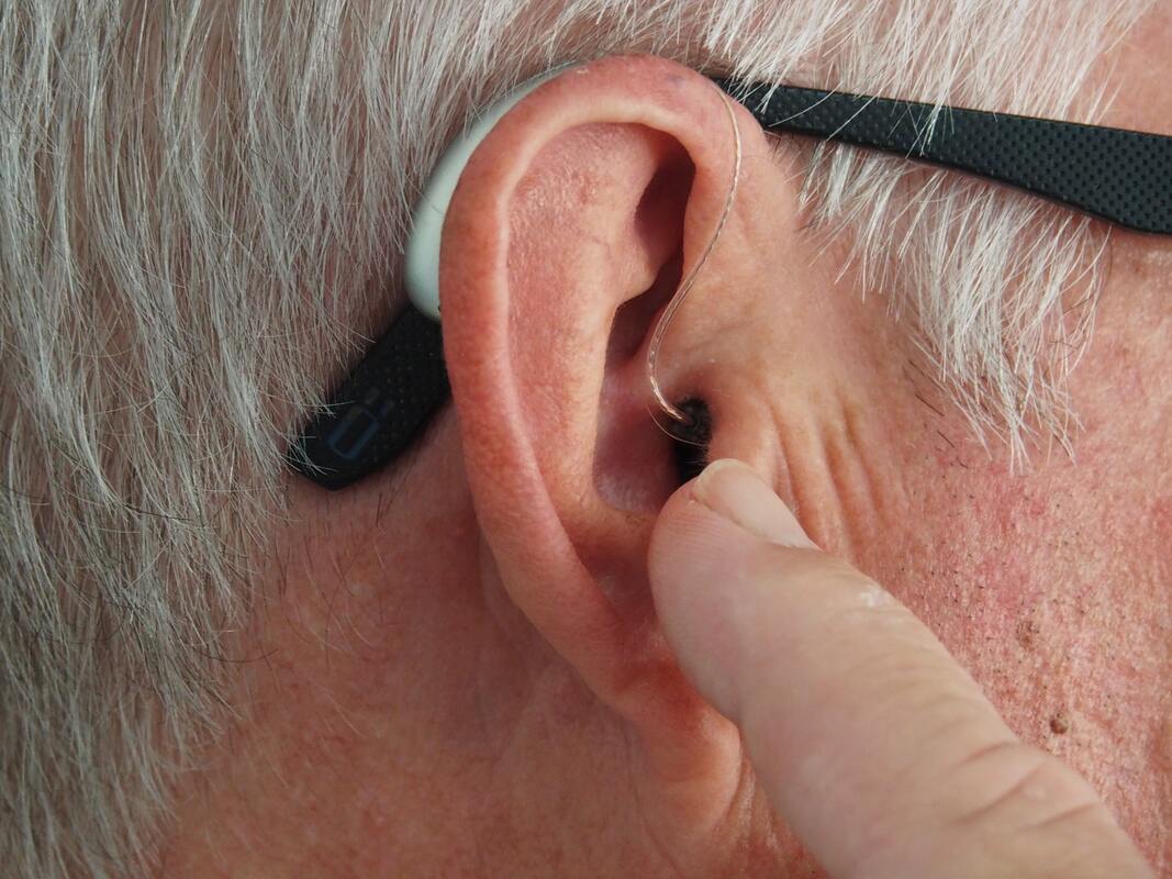 man_white_hair_finger_pointing_at_ear_and_receiver_with_silver_BTE_hearing_aid