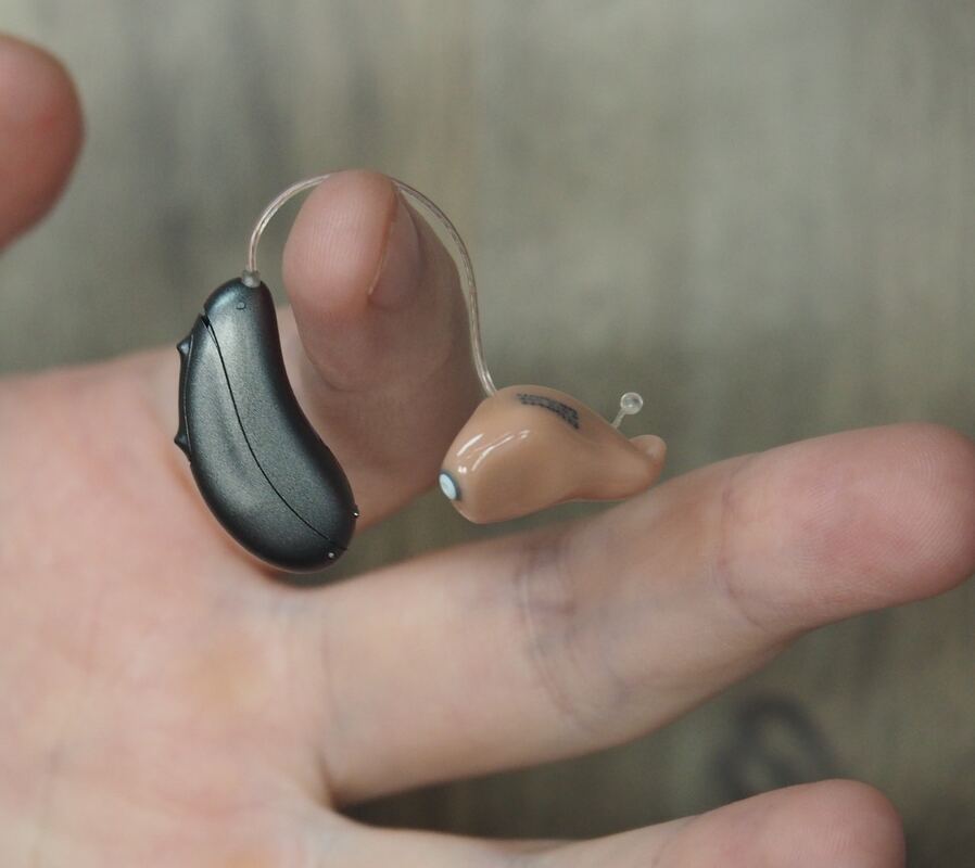 A hand holds a black behind-the-ear hearing aid with a customized receiver for the ear.