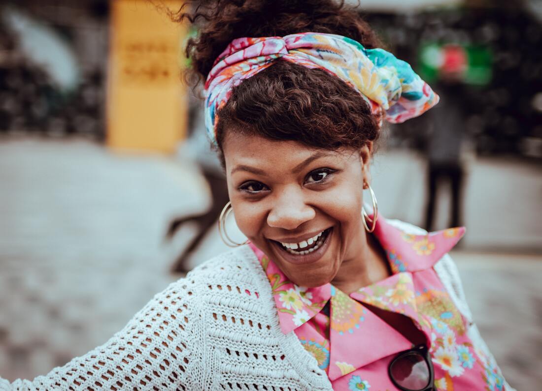 A D/deaf woman smiles, wearing a multi-colored headscarf, a pink floral blouse, and a white sweater.