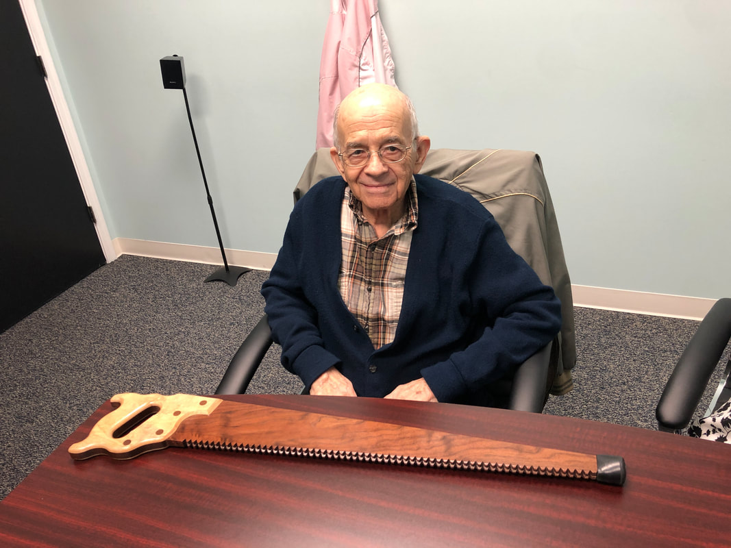 elderly man with glasses and hearing aids wears navy blue sweater and grey plaid shirt while sitting in lititz hearing aid office with brown walking cane on desk shaped like a hand saw