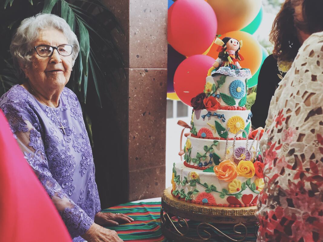 An older woman with hearing aids and glasses celebrates her birthday with a floral-decorated cake and doll cake topper.