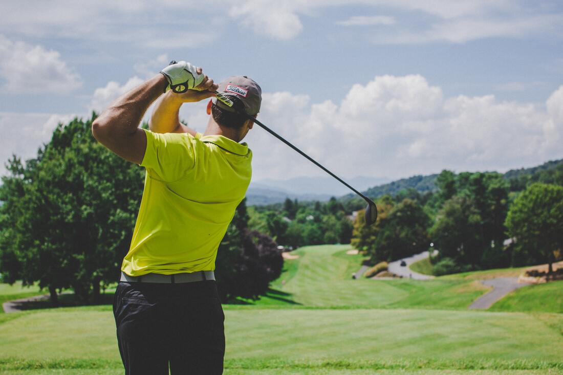 A golfer in a neon yellow shirt, black pants, and a grey baseball cap swings a golf club on a green golf course.