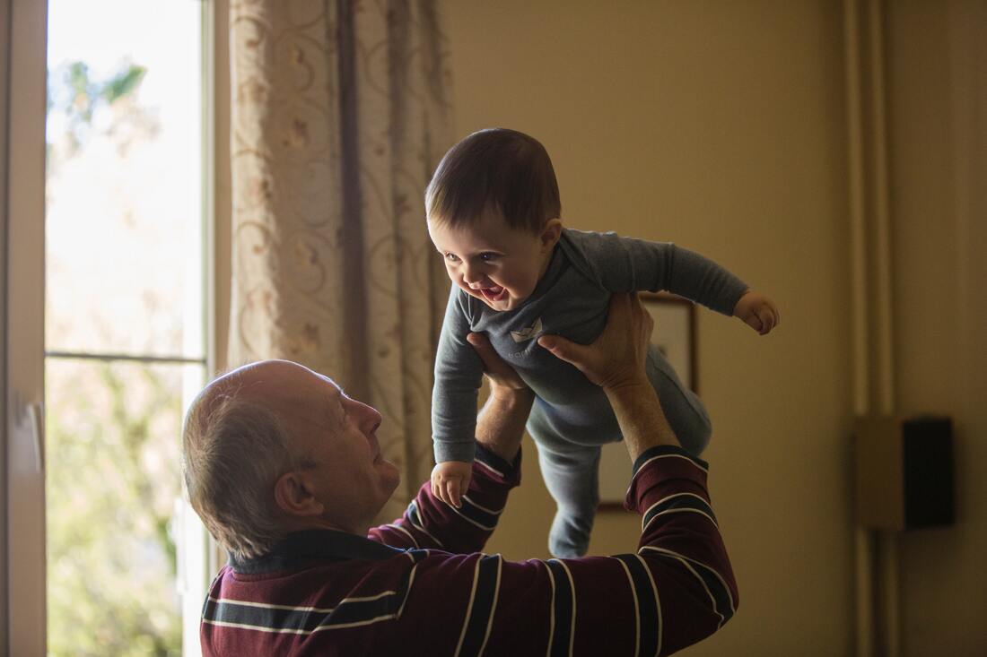 A man with hearing loss wears a maroon, white, and blue shirt and holds a smiling grandchild in a bluish-grey onesie.