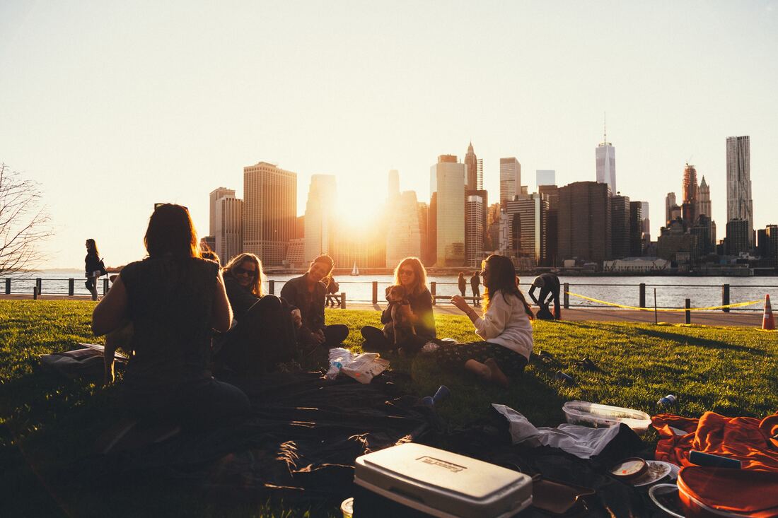 A group of friends talk and smile while sitting outside in the grass with a city skyline in the background.