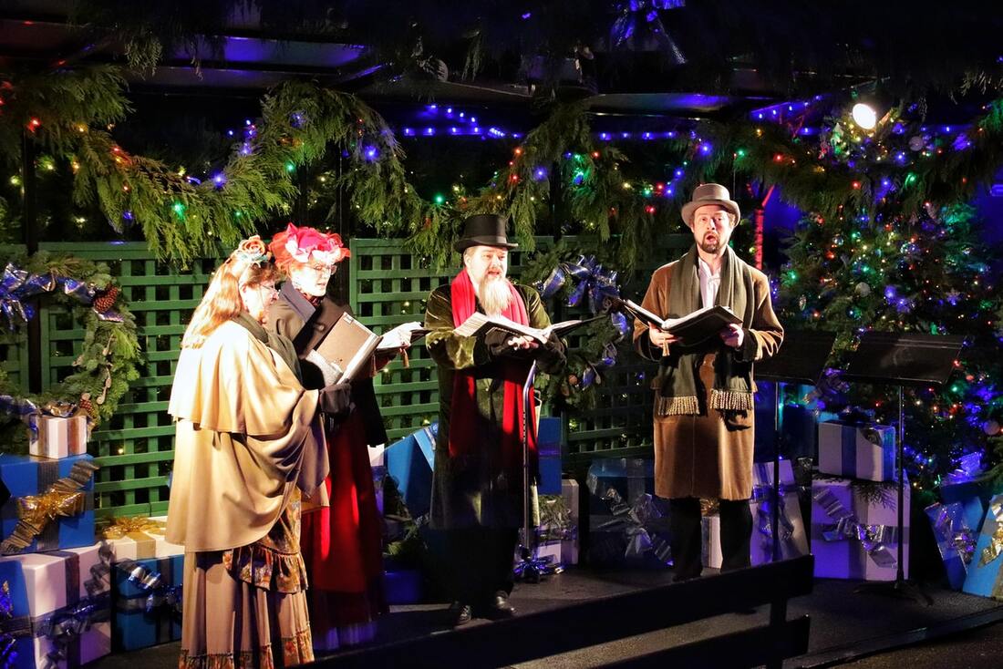 Carolers dressed up in old-timey clothes sing in front of a Christmas display. 