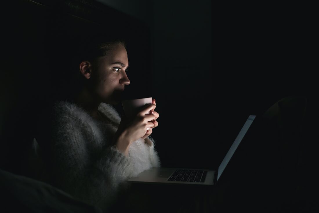 A woman sits in the dark while holding a cup to her mouth and watches a scary movie from a glowing laptop.