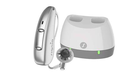 A silver behind-the-ear hearing aid featuring a white and grey charger port with green lights.