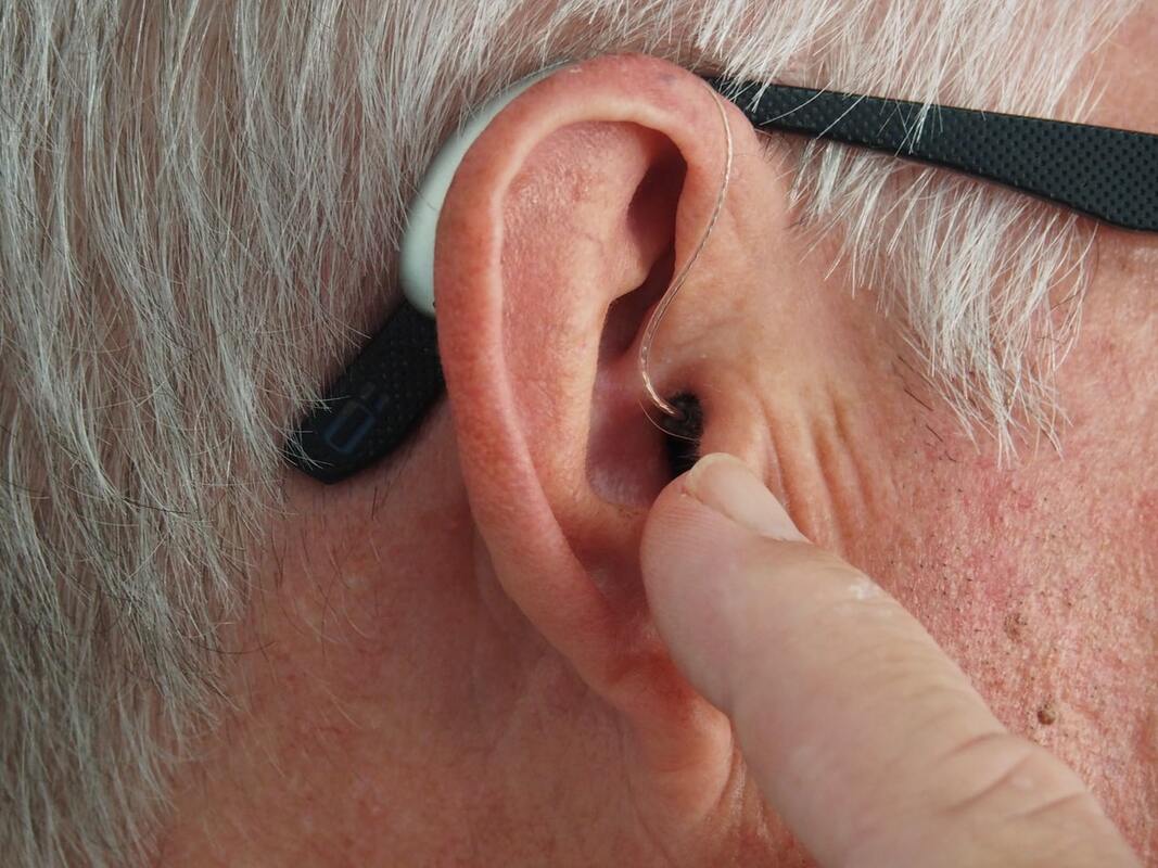 A man with white hair and eyeglasses points his finger at his ear and receiver with a silver behind-the-ear hearing aid.