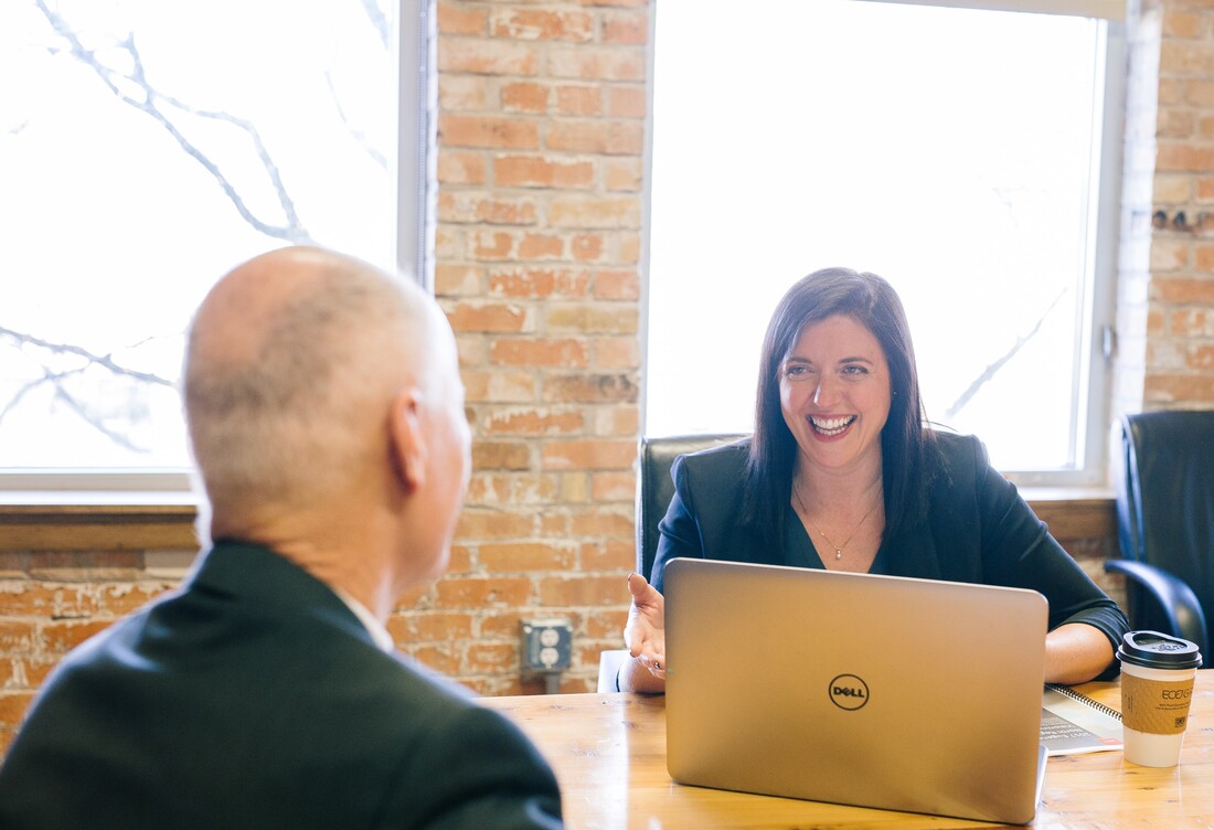 A woman with a silver laptop smiles at a man in an office during his hearing aid consultation.