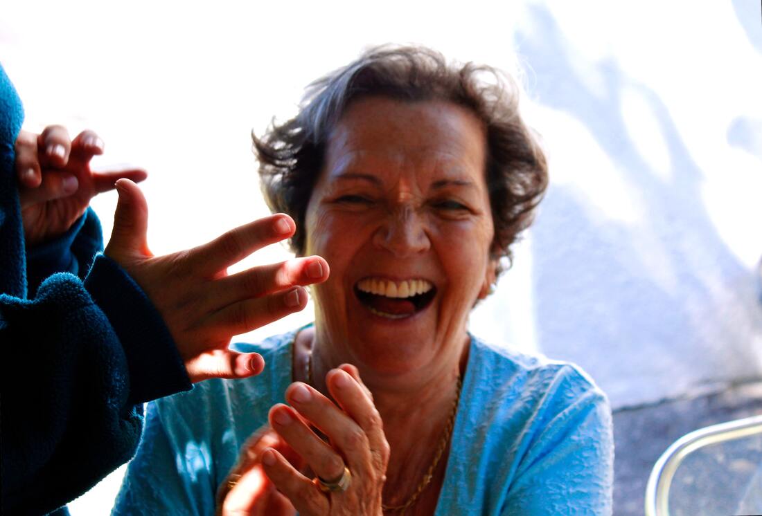 A woman with grey hair and a blue shirt laughs and smiles while clapping her hands.