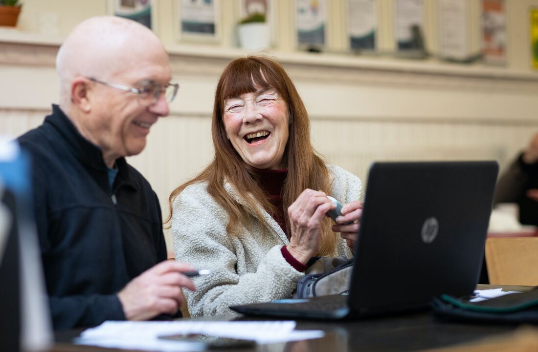 An older man with hearing aids and a woman look at a laptop and laugh.
