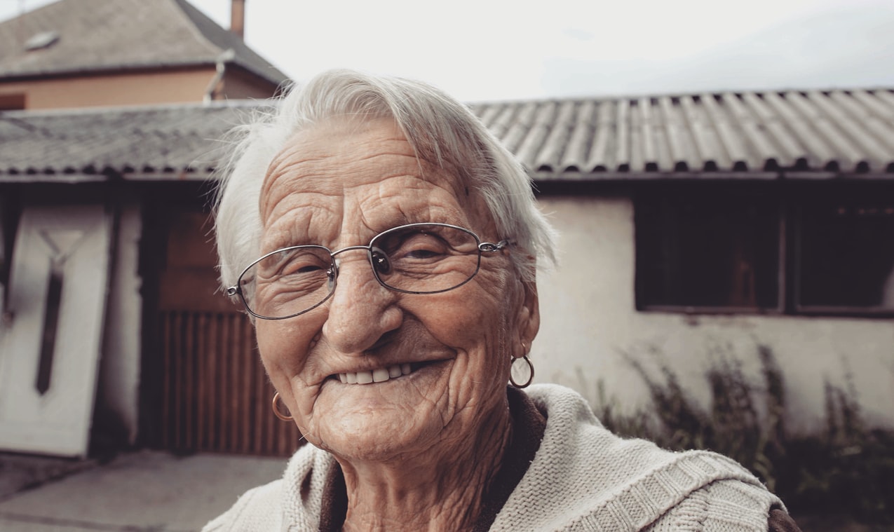woman_in_front_of_white_home_with_tin_roof_has_wrinkles,_white_hair,_glasses_hoop_earrings_hears_better_with_hearing_aids