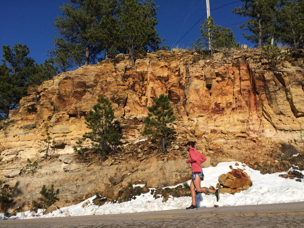 A man jogs by a rocky mountain with trees at the top and snow on the side of the road.
