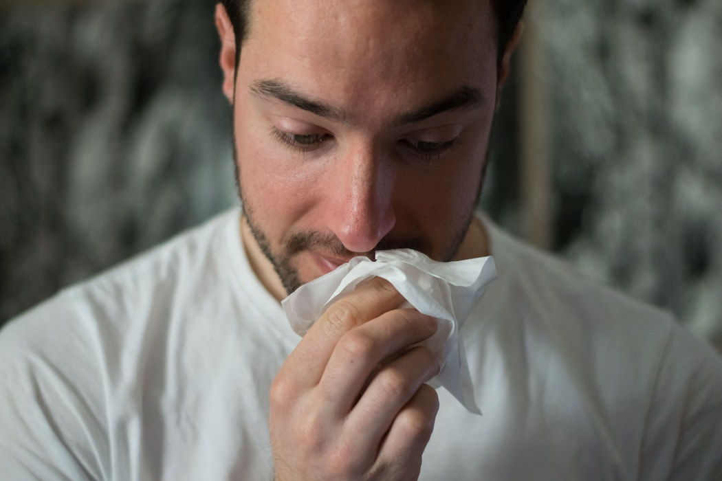 A man with a mustache and beard wears a white t-shirt and holds a tissue under his nose while looking down.