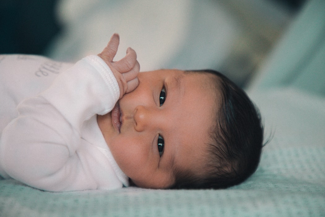 A newborn baby with dark hair and dark eyes lies down and looks straight ahead with hand on cheek.