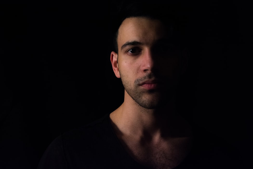 A man stands in a shadowy black background with half of his face exposed to the light.