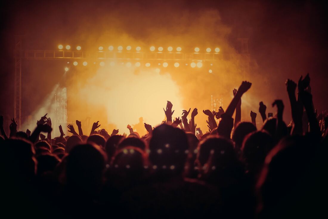 A crowd of concertgoers wave their hands and watch a live band’s performance on a stage bathed in orange-yellow lights.