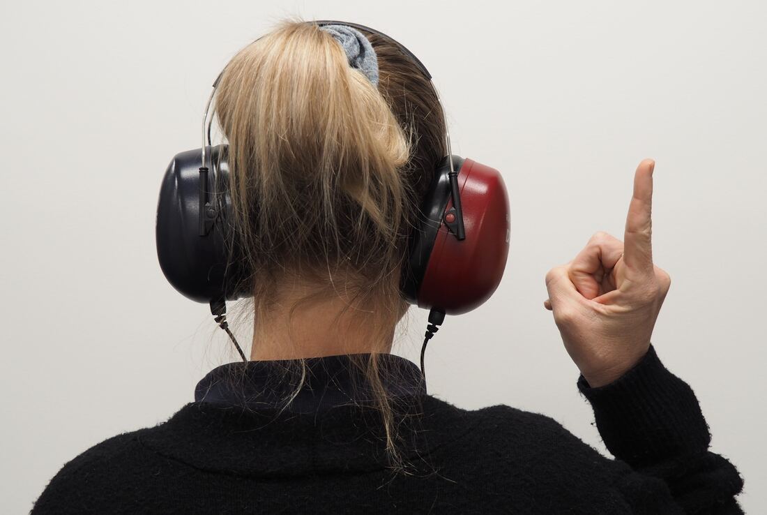 A woman wearing a black sweater wears black and red headphones while pointing her finger up during a hearing test.