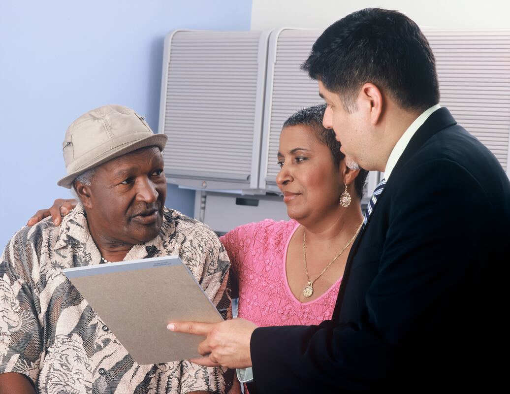 An older man wearing a tan shirt and bucket hat and his wife receive a consultation with a hearing aid provider in Lancaster.
