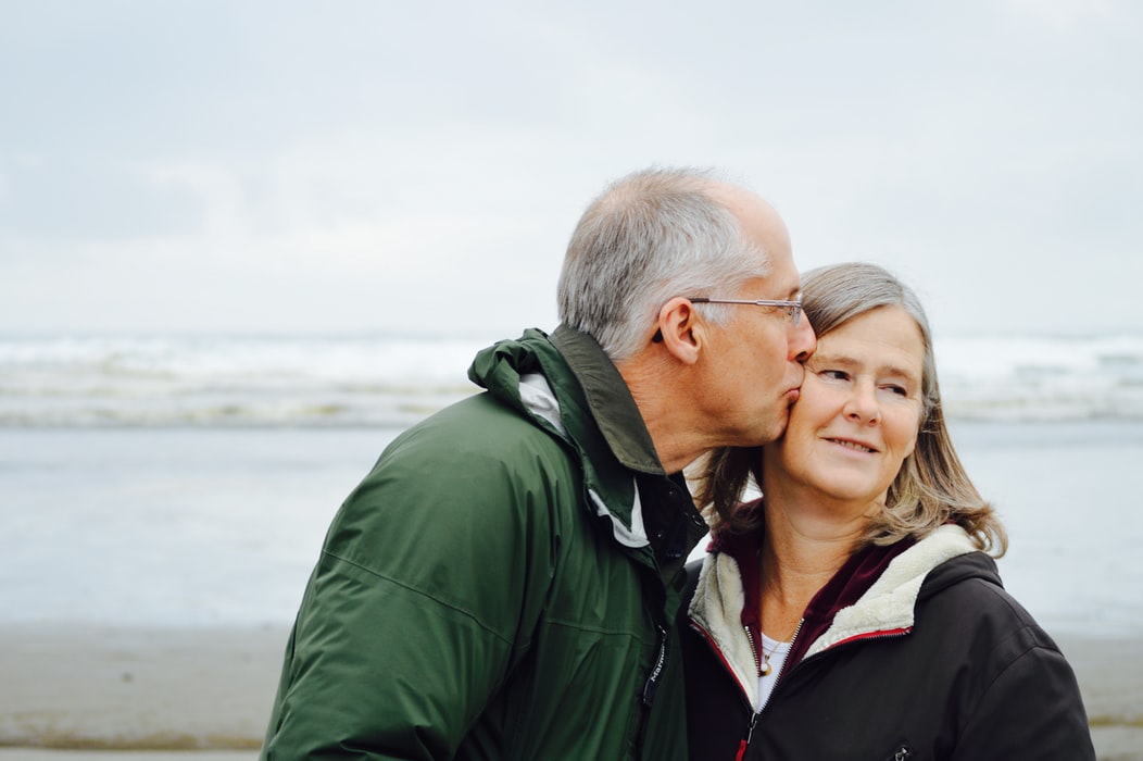 A man with hearing loss, glasses, and a green jacket kisses a woman on the cheek by the ocean.