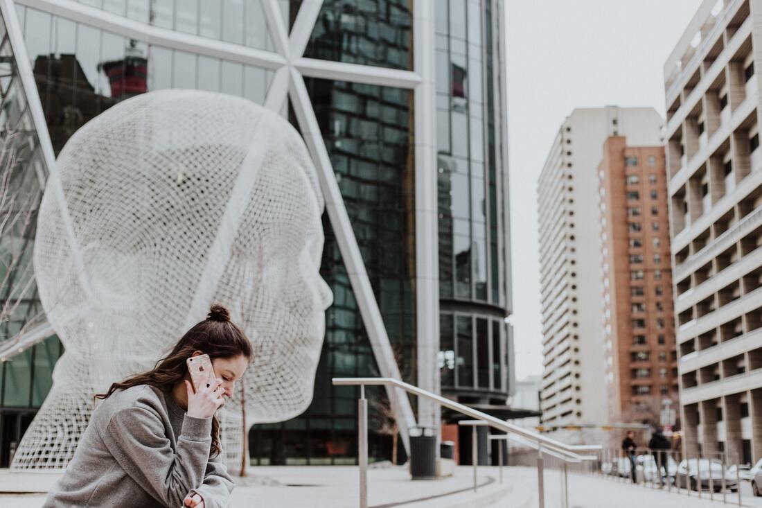 A woman with hearing loss listens and talks on her phone near a large white sculpture in a city.