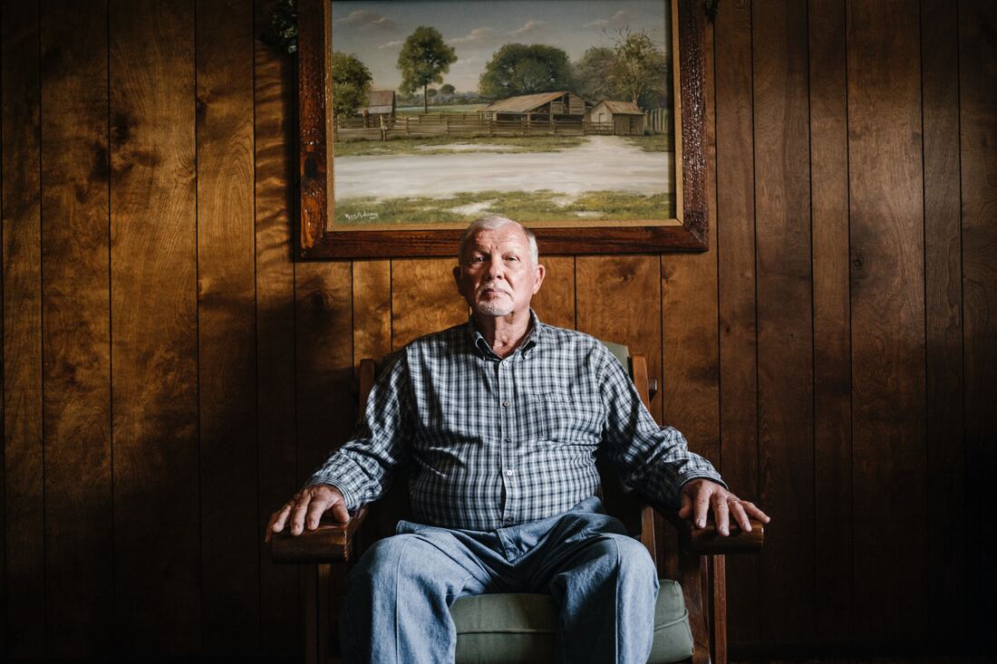 lonely old man with hearing loss wearing plaid shirt and jeans sits in chair under painting of barn on farm hanging on wood panelled wall in lancaster county