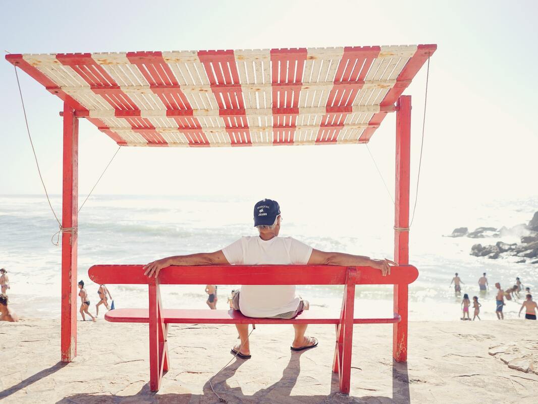 man at beach sits on red bench with red and white roof
