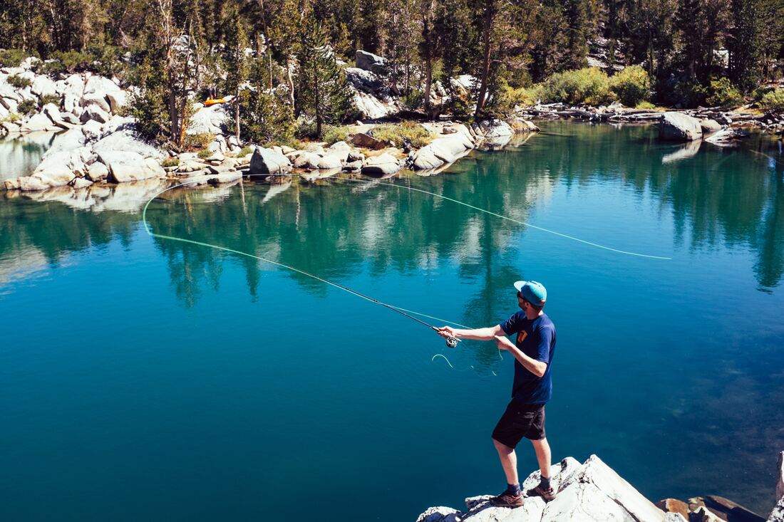 A man stands on a rock by a quiet lake and casts a fishing reel.