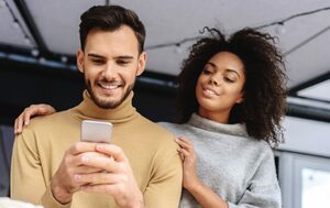 man with beard in yellow turtleneck holds smartphone connected to hearing aids with bluetooth and woman with black hair and grey turtleneck sweater stands by