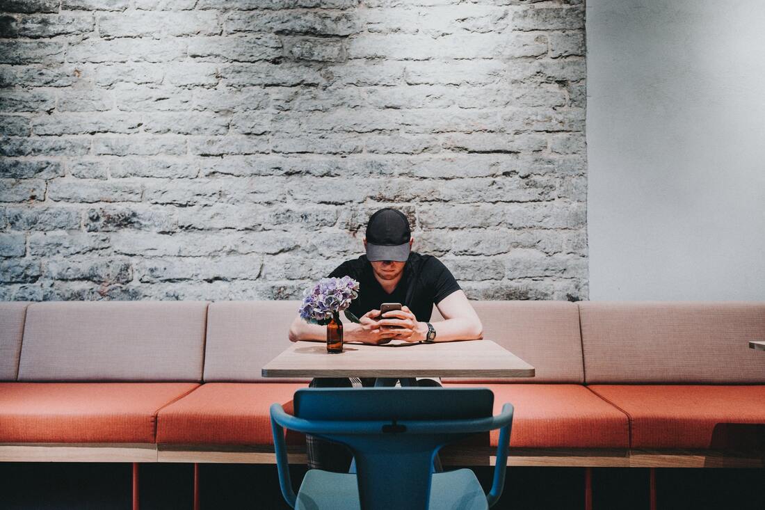 man with black baseball cap and black t-shirt sits alone in public space and looks at phone