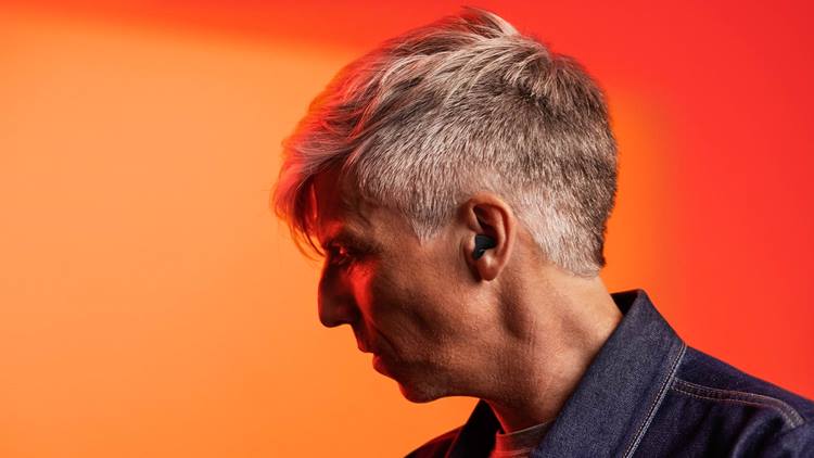 man with black rechargeable signia hearing aid in ear wears jean shirt and stands in front of orange and red backdrop