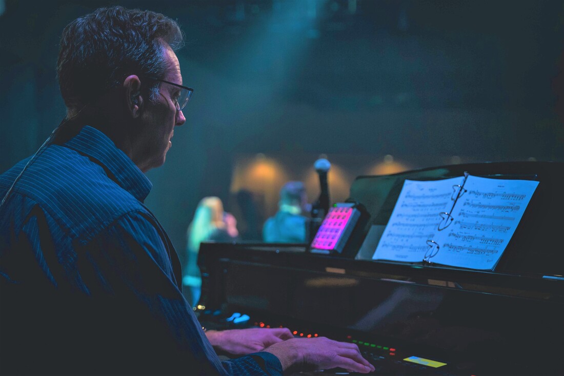 A man with hearing aids plays the piano while looking at music sheets under multi-colored stage lights.