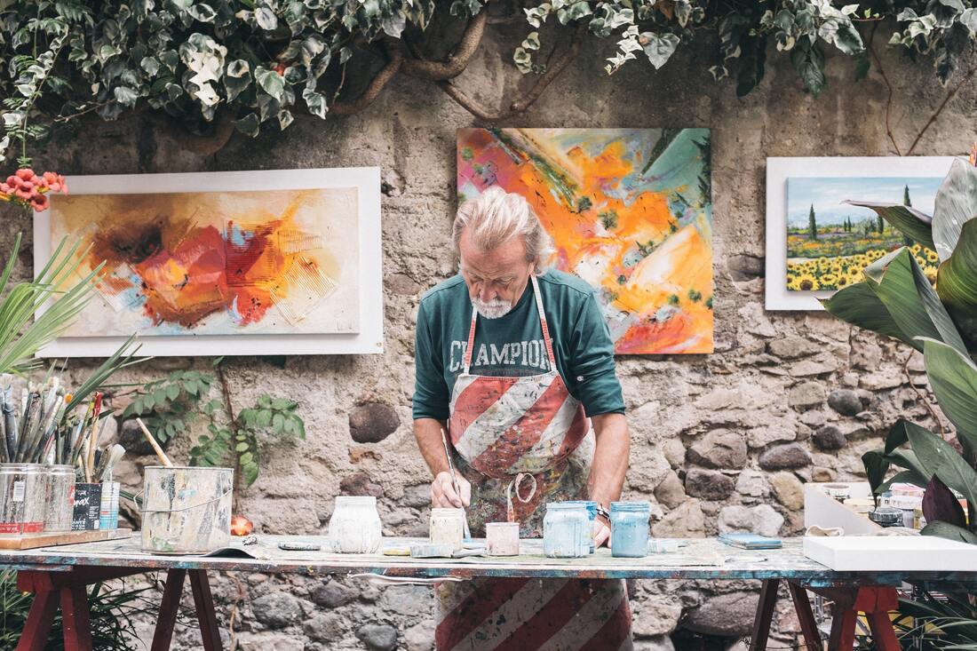 man with hearing loss has art supplies paints with brush and paintings hanging in background