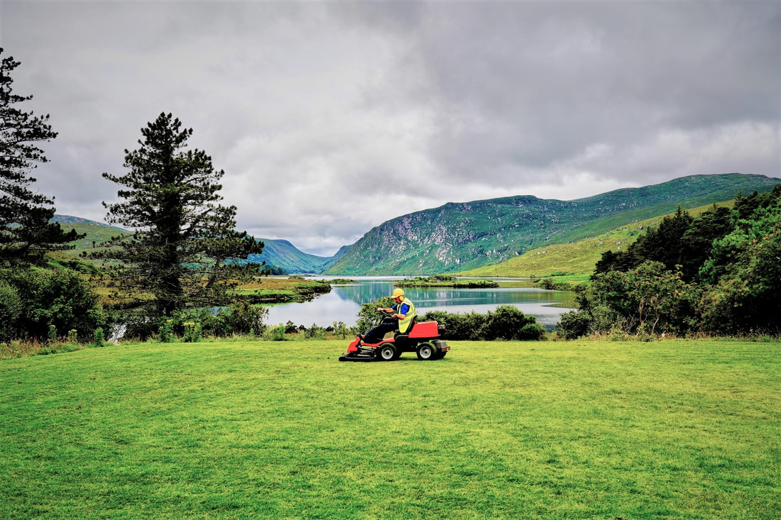 man with helmet earmuffs and yellow reflection gear drives loud gas powered lawn mower on grass by the lake 