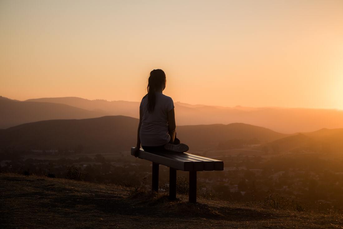 A woman sits on a bench watching the sunset while practicing listening exercises and meditating.