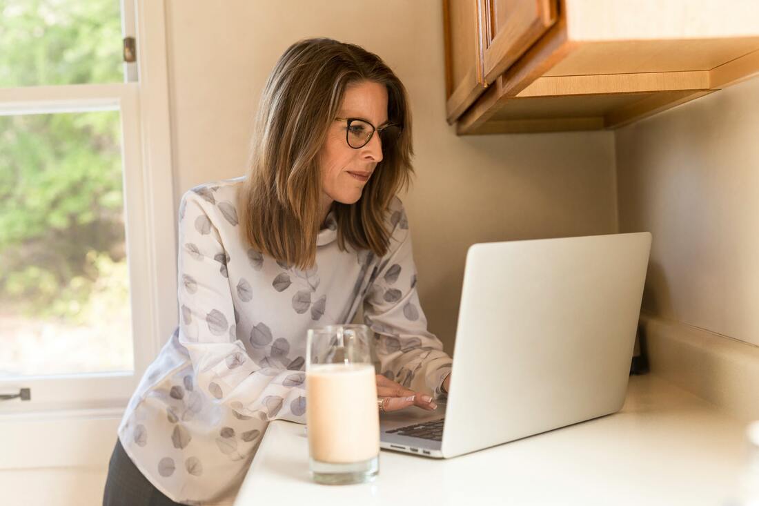 A menopausal woman with glasses looks at hearing loss information on a laptop in her kitchen, with glass of milk by her side.