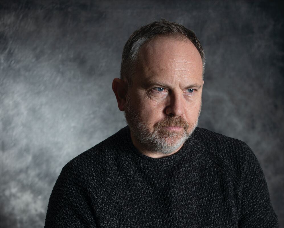 A man with blue eyes and a graying beard wears a black sweater and looks off in the distance.