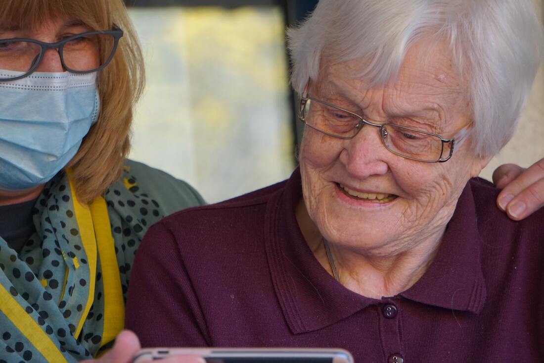 A woman wearing a blue facemask and glasses stands by a woman with white hair and eyeglasses while they look at a smartphone.