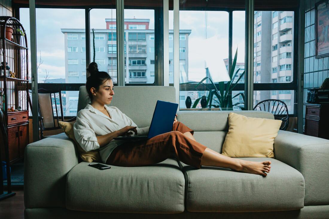 A woman with hearing aids sits on a grey couch with a blue laptop in an apartment by a glass window.
